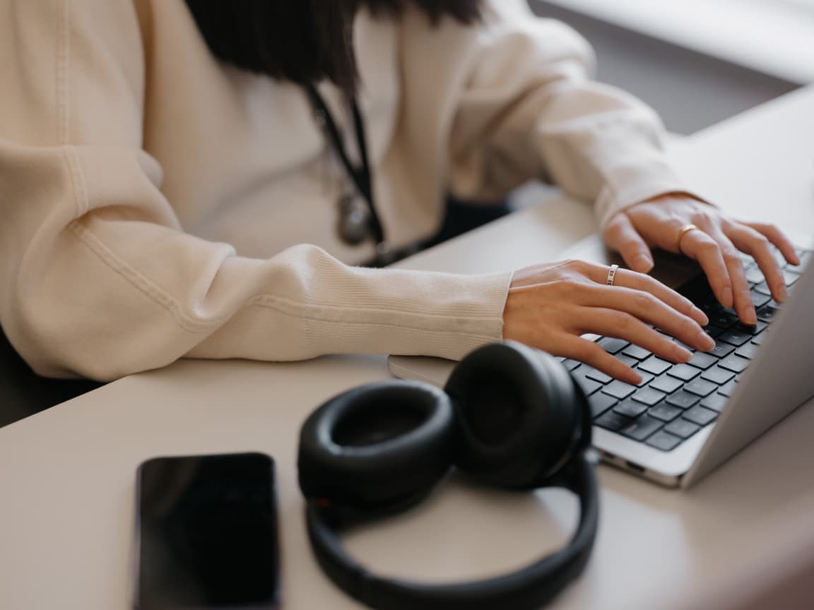 A person in a cream sweater types on a laptop, with a pair of black headphones and a phone on the desk beside it.